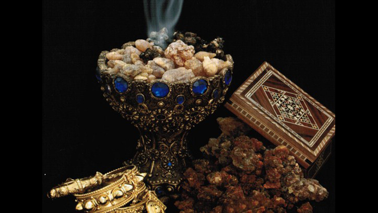 golden chalice with blue gems around the lip filled with smoldering frankencense a decorative box open with myrrh falling out and pieces of gold bracelets
